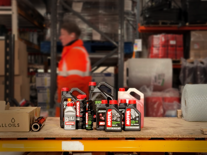 Collection of Motul oil bottles on a table in a warehouse setting with a person in an orange safety vest in the background.